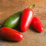 A group of jalapeño peppers on a stone surface, with one green and three having turned red, showcases different ripeness stages. The background features a textured, earthy-toned tile, illustrating what to do as your jalapeños transition in color.