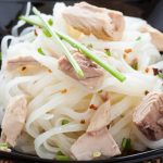 A black square dish filled with rice vermicelli topped with chunks of chicken, garnished with sliced scallions and red pepper flakes. The healthy meal is elegantly set on a bamboo placemat.