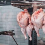 Two whole, raw chickens with glistening skin hang by metal hooks in a glass display case, contrasting against the white tiled wall in the background.