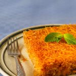 A plate with a piece of golden-brown kunafa, woven with delicate vermicelli and topped with mint leaves. A fork rests on the plate against a blue checkered tablecloth.