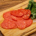 Slices of gluten-free pepperoni are artfully arranged on a wooden cutting board, accompanied by vibrant kale leaves. The board rests elegantly on a rustic wooden surface.