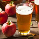 A glass of amber apple cider labeled Loon Juice sits on a wooden table surrounded by fresh red apples. Another glass is partially visible in the background. A wicker basket is filled with more apples, completing a rustic and refreshing scene that answers all cravings for natural simplicity.