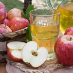 Fresh apples in a wicker basket sit beside a wooden board with sliced apples and a glass of gluten-free apple juice. A pitcher of juice and whole apples complete the natural, outdoor setting, conveying a sense of freshness and harvest.