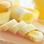Close-up of a sliced banana on a wooden cutting board. The slices, perfect for storage or a quick snack, are neatly arranged, with the unpeeled fruit and its peel visible in the background. The focus is on the fresh, yellow banana pieces, ready to enjoy or store in the fridge for later.