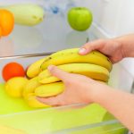 A person carefully places ripening bananas into a refrigerator drawer, hoping they'll last longer. Nearby, oranges, a green apple, and a zucchini are neatly arranged on the shelf and in the drawer.