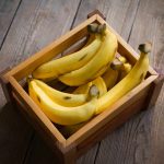 A wooden crate filled with ripe yellow bananas sits on a rustic kitchen counter. The bananas, adorned with small brown spots, indicate their perfect ripeness. The crate's simple design contrasts beautifully with the textured wood flooring.
