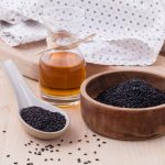 A wooden bowl filled with black sesame seeds sits beside a small ceramic spoon also containing seeds, showcasing the essence of gluten-free ingredients. A glass bottle of rich sesame oil is next to the bowl, and a white cloth with a subtle pattern lies in the background on a wooden surface.