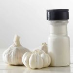 A jar of garlic powder with a black lid stands on a white surface, proudly displaying its gluten-free promise. Two whole garlic bulbs rest beside it, enhancing the pure essence. The backdrop is a soft, blurry white, emphasizing the product’s natural and wholesome appeal.