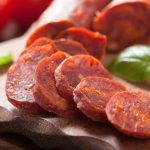 Sliced chorizo on a wooden cutting board, with fresh basil leaves beside it. The rich red color and textured surface come alive, enhanced by sodium lactate. In the background, whole sausages and a hint of tomatoes add depth to this appetizing scene. Always check for allergen info before indulging.