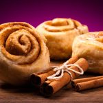 Four Pillsbury cinnamon rolls are arranged on a wooden surface with three cinnamon sticks tied together in front. The background is a rich purple, enhancing the golden brown color of these tempting pastries.