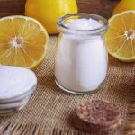 A small glass jar filled with gluten-free white powder is placed on a burlap surface, surrounded by halved and whole lemons. A small bowl containing similar citric acid powder is nearby, with a cork lid lying beside it.