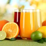 Two glasses of vibrant fruit juices, one orange and one red, are surrounded by whole and sliced citrus fruits like oranges, lemons, and limes. The softly blurred background suggests a sunny outdoor setting that's perfect for enjoying these gluten-free treats.