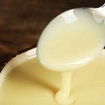 A spoon drizzles creamy, gluten-free condensed milk over a bowl, with a close-up view highlighting the milk's thick texture and glossy surface on a wooden background.