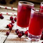 Glasses of gluten-free cranberry juice are placed on a rustic wooden table, surrounded by scattered fresh cranberries. Straws with red polka dots lie in the background, adding a festive touch to the scene.