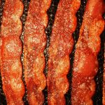 Close-up of five sizzling strips of crispy bacon cooking in a pan. The bacon is golden brown and glistens with hot oil, enticingly cooked to perfection.
