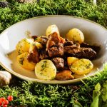 A plate of roasted elk meat and mushrooms served with boiled potatoes garnished with herbs, surrounded by a bed of fresh greenery and scattered brown mushrooms.