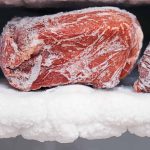 Close-up of three pieces of raw venison, covered in frost, resting on a thick layer of ice inside cold storage. The surface of the meat reveals intricate white frost patterns, emphasizing the chill within the freezer.