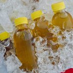Four bottles with yellow caps filled with brown liquid labeled Juicy Juice are submerged in crushed ice, likely to keep them cool. The setting suggests an outdoor market or refreshment stall, leaving questions about their unique flavors eagerly answered by curious passersby.