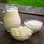 A glass jug of milk, a bowl of cottage cheese, and a bowl of yogurt made with lactic acid starter cultures rest on a rustic wooden table. The setting is enhanced by the vibrant green background, evoking the freshness of an outdoor dairy scene.