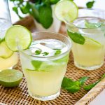 Three glasses of refreshing lime mojitos sit on a wicker tray, made with different types of lime juices. Each glass is garnished with a lime slice and mint leaves. A halved lime and a plant are in the background, adding a fresh and vibrant touch. Plus, they're gluten-free!