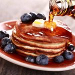 A friendly stack of pancakes with butter on top, drizzled with rich maple syrup, is surrounded by fresh blueberries on a plate.