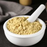 A white mortar and pestle filled with light brown ginger powder is placed on a dark surface. A striped cloth is folded in the background, and a metal scoop with more mustard powder is partially visible on the right, ensuring this gluten-free ingredient is ready for use.