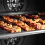 Slices of chewy bacon sizzle on a tray inside the oven. The crispy strips are evenly spaced, while the warm glow of the stove lights up the background.
