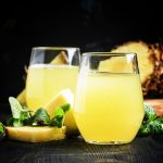 Two glasses of gluten-free pineapple juice sit on a wooden table, with fresh pineapple pieces and mint leaves scattered around. The dark background highlights the vibrant yellow juice and the fresh green mint, telling a story of refreshing indulgence.