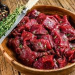A wooden bowl filled with raw venison, garnished with fresh herbs, sits on a rustic wooden table. A knife, adorned with a sprig of herbs on the handle, rests atop the bowl. In the background, a small dish holds a colorful mix of peppercorns.