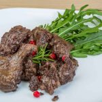 A white plate with perfectly cooked venison roast and liver pieces, artfully garnished with fresh arugula, rosemary, and red berries, sits invitingly on a wooden table.