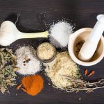 A collection of spices and herbs on a dark wooden surface includes a bulb of garlic, gluten-free coarse salt, leafy herbs, red pepper flakes, ground spices, and a mortar and pestle with seeds.