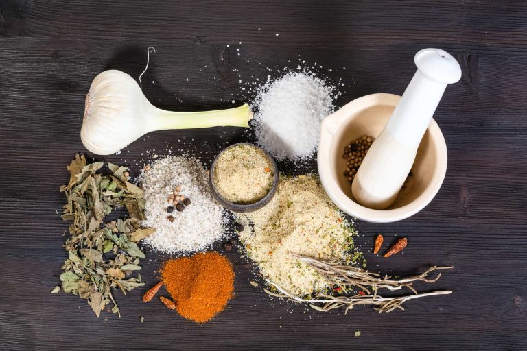 A collection of spices and herbs on a dark wooden surface includes a bulb of garlic, gluten-free coarse salt, leafy herbs, red pepper flakes, ground spices, and a mortar and pestle with seeds.