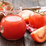 A glass pitcher filled with refreshing, gluten-free tomato juice sits on a wooden surface. Next to it, a red and white checkered cloth cradles halved and whole tomatoes. A basket brimming with more tomatoes graces the scene in the background.