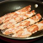 Slices of bacon sizzling in a black frying pan are partially cooked, some areas crispy and others still chewy. Small bubbles of oil appear around the meat, ensuring each piece is safe to eat. The pan rests steadily on a stovetop, tempting with its aroma.