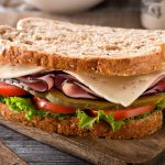 A hearty sandwich on a wooden board, featuring gluten-free bread, layers of ham, Swiss cheese, fresh lettuce, tomato slices, and pickles. A knife lies nearby on the rustic wooden surface.