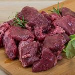 Raw venison chunks on a wooden cutting board, garnished with sprigs of rosemary. Nearby are a bay leaf, a fresh red tomato, and leafy greens, ready to be chilled in the fridge to lock in freshness.
