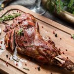 Roasted lamb leg on a wooden cutting board, garnished with rosemary and peppercorns, with cutlery beside it. The rustic presentation, surrounded by greenery, mirrors the earthy allure of venison taste and invites comparisons to game dishes in both aroma and texture.