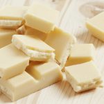 Chunks of gluten-free white chocolate are stacked randomly on a light wooden surface. The texture is smooth and slightly crumbly at the edges, reflecting light gently. The background showcases a natural wood grain pattern, enhancing the luxurious appeal of this sweet indulgence.