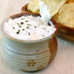 A rustic ceramic bowl filled with creamy, gluten-free dip, garnished with chopped chives and made with rich sour cream, features a tortilla chip dipped in. In the background, a wooden bowl holds several tortilla chips on a neutral-colored surface.