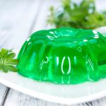 A bright green Jelly Ace dessert on a white plate, garnished with a small sprig of greenery. The gelatin is molded into a ring shape with a smooth, glossy surface. Additional greenery is blurred in the background on a light wooden table.