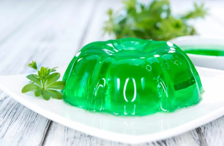 A bright green Jelly Ace dessert on a white plate, garnished with a small sprig of greenery. The gelatin is molded into a ring shape with a smooth, glossy surface. Additional greenery is blurred in the background on a light wooden table.