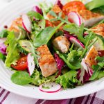 A colorful salad with grilled chicken, cherry tomatoes, sliced radishes, cucumber, and mixed greens on a white plate meets the highest food standards. The plate rests on a red and white striped cloth.