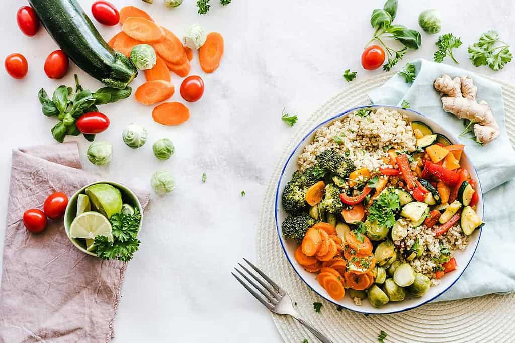 A colorful bowl of quinoa salad with broccoli, carrots, zucchini, and red peppers sits on a circular mat. Perfect for paleo and vegan recipes, fresh vegetables, lime slices, and a fork surround the bowl on a light surface.