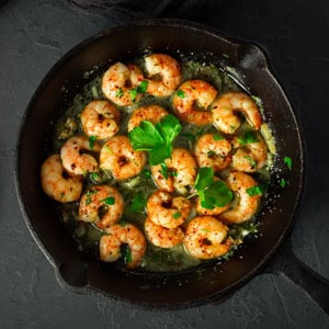 kitchenware A skillet filled with cooked shrimp in a garlic butter sauce, reminiscent of paleo indulgence, garnished with fresh parsley. The shrimp are seasoned with pepper and arranged neatly, creating an appetizing presentation on a dark background.