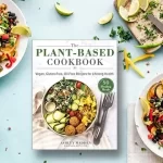 A cookbook titled "The Plant-Based CookBook" rests on a blue surface, encircled by vibrant salads featuring black beans, avocado, tomatoes, and lime wedges. Plates and bowls showcase these colorful creations with utensils and garnish thoughtfully scattered around.