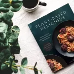 A vegan cookbook titled "Plant-Based Delicious" lies open on a table next to a leafy plant and a cup of coffee. The cover showcases a plate of spaghetti with sauce and meatballs, perfectly highlighting its plant-based twist.