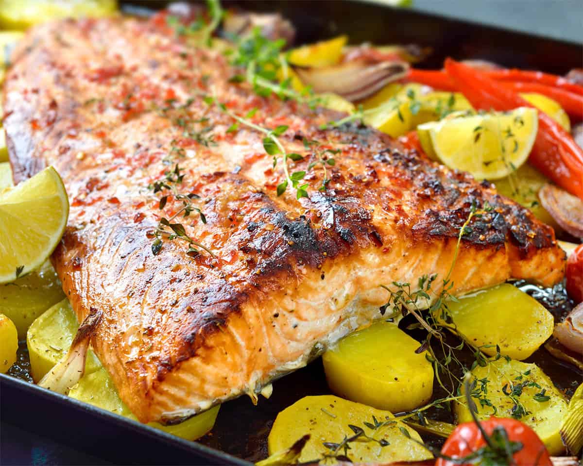 Paleo grilled salmon skin fillet garnished with herbs rests on a bed of roasted potatoes, cherry tomatoes, and lemon slices, crafting a vibrant and appetizing dish.