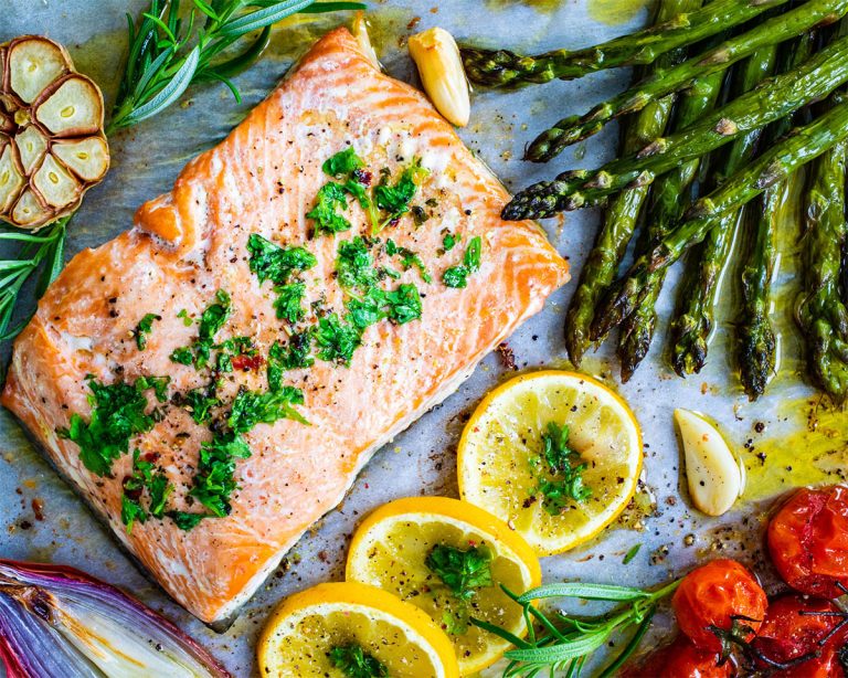 A paleo-friendly baked salmon fillet, garnished with herbs, is nestled among roasted asparagus, cherry tomatoes, lemon slices, garlic cloves, and rosemary sprigs on a baking sheet.