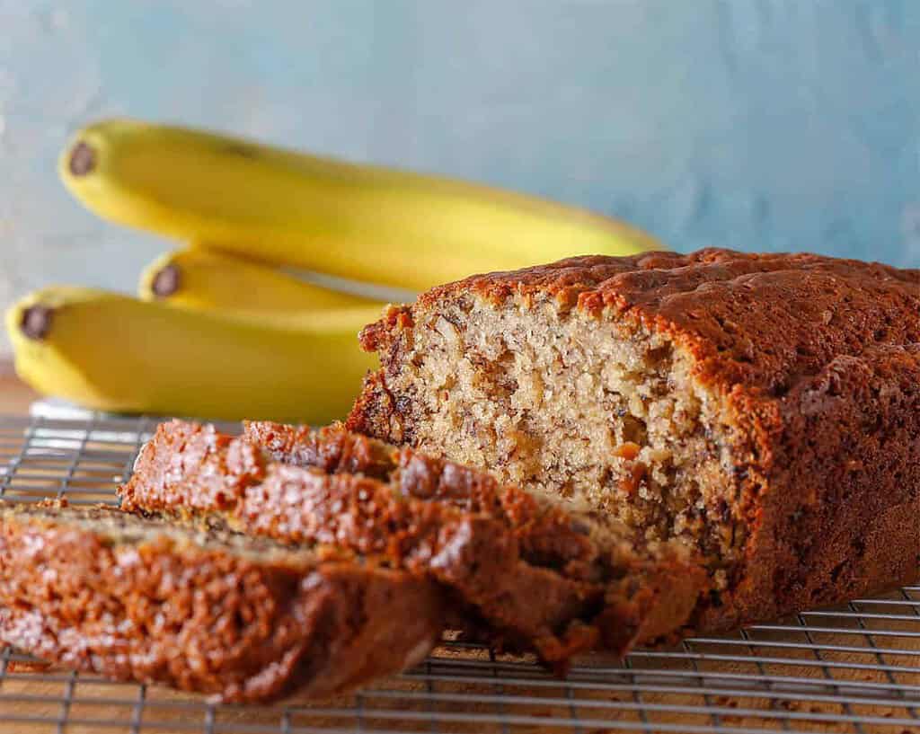 A loaf of paleo banana bread is sliced on a cooling rack, with three bananas in the background against a light blue textured wall. The bread boasts a golden-brown crust and a moist, dense interior, showcasing chunks of ripe banana.