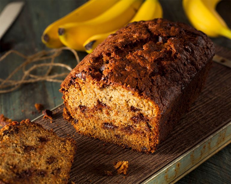 A loaf of Paleo banana bread with chocolate chips is sliced on a wooden board. Two bananas are in the background, and a piece of twine is visible. The bread has a golden-brown crust and a moist interior, making it the perfect recipe for those seeking deliciously healthy indulgence.